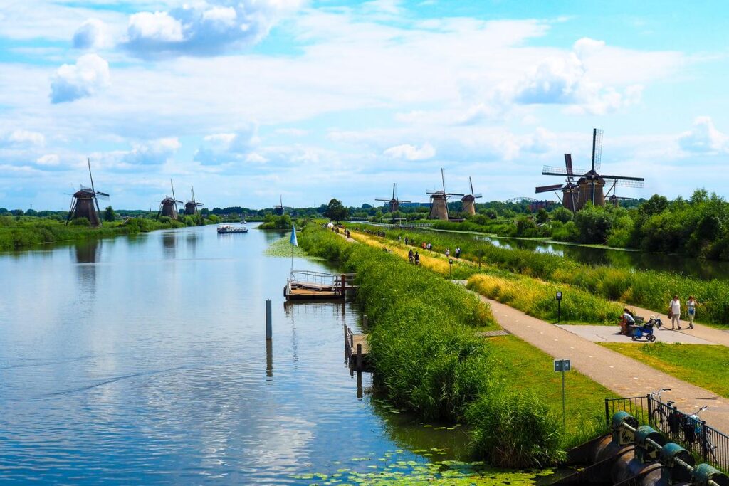 A sunny day at Kinderdijk featuring historic Dutch windmills, a boat on the water, and visitors walking along a lush green path.