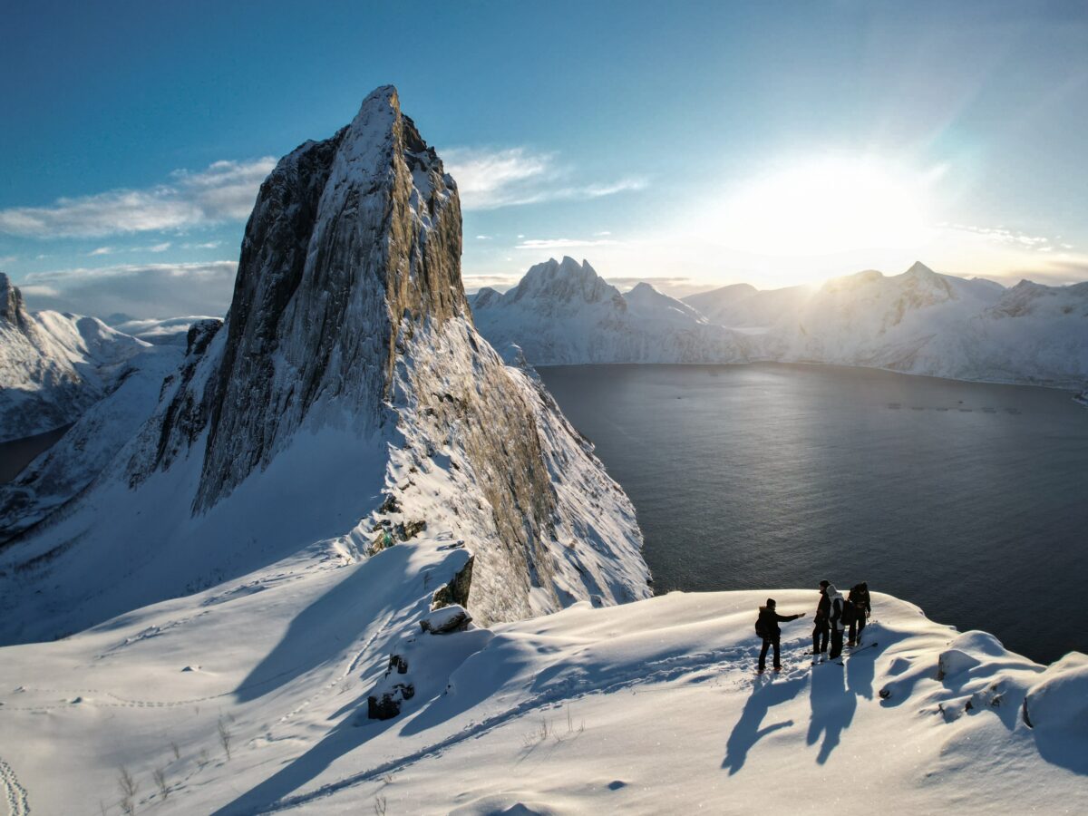 Snowshoe hikers on a snowy ridge with Segla Mountain, Norway, overlooking a calm Arctic fjord.