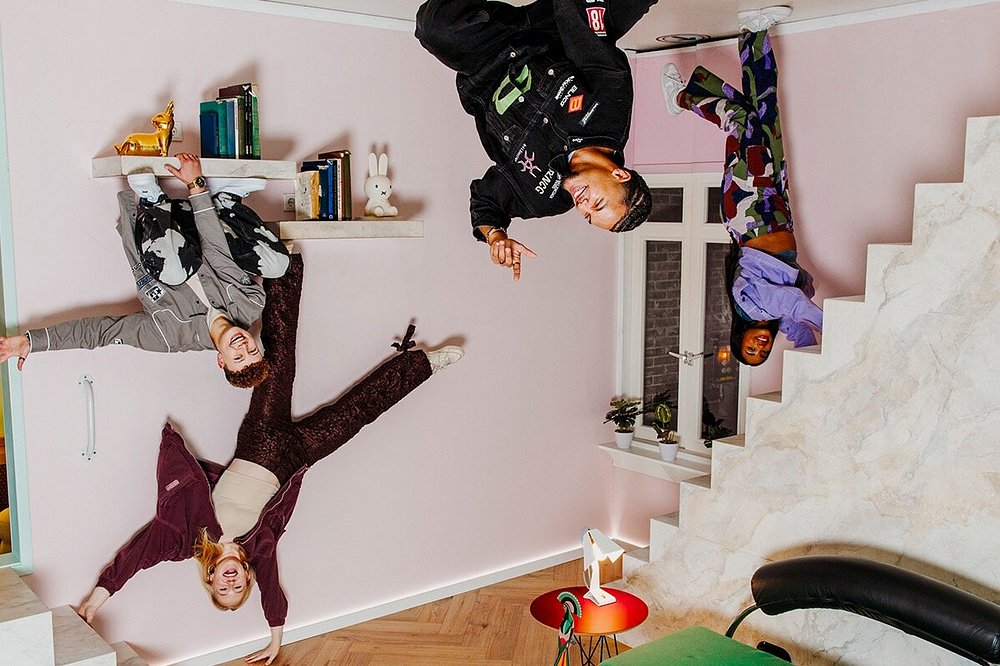 Playful surreal interior with four young adults defying gravity amid pastel walls and eclectic decor.