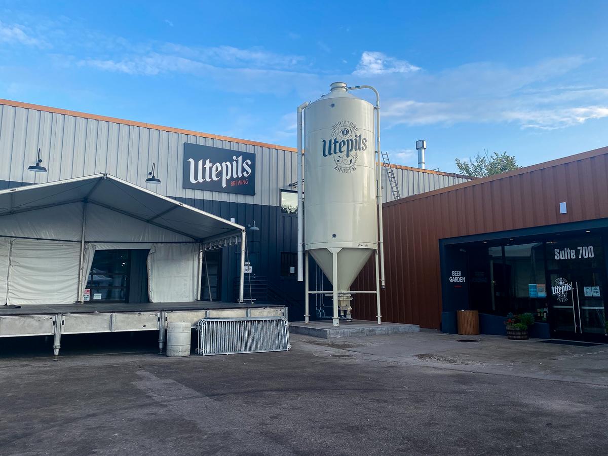 Sunny view of Utepils Brewing's facade with branded beer silo, outdoor seating area under a tent, and entrance to beer garden in Minneapolis, Minnesota.