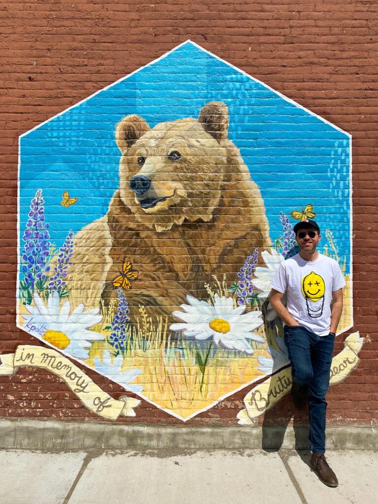Kyle Kroeger, writer posing with Brutus bear mural in Livingston, Montana showcasing wildlife and wildflowers.