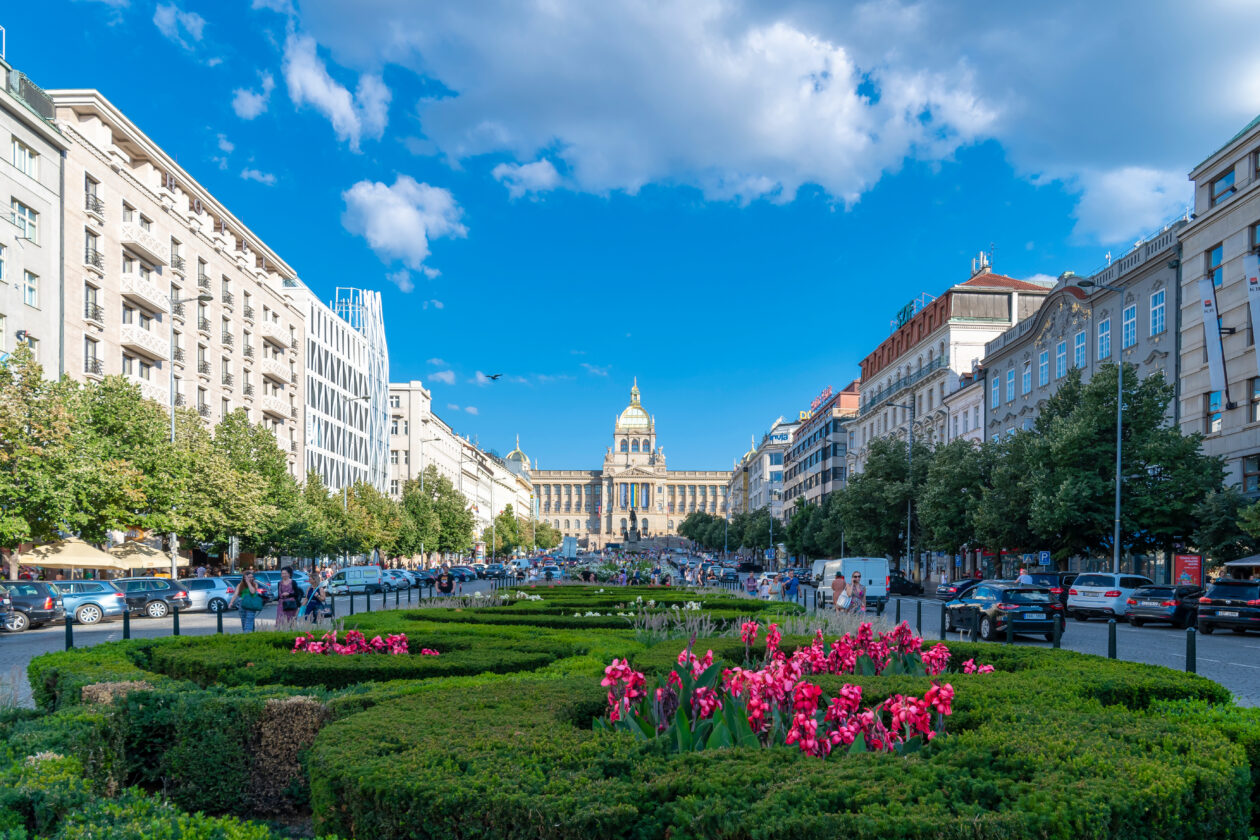 Pragues Wenceslas Square view: National Museum, ornate façades, pink blossoms, trees, urban life in Bohemia.