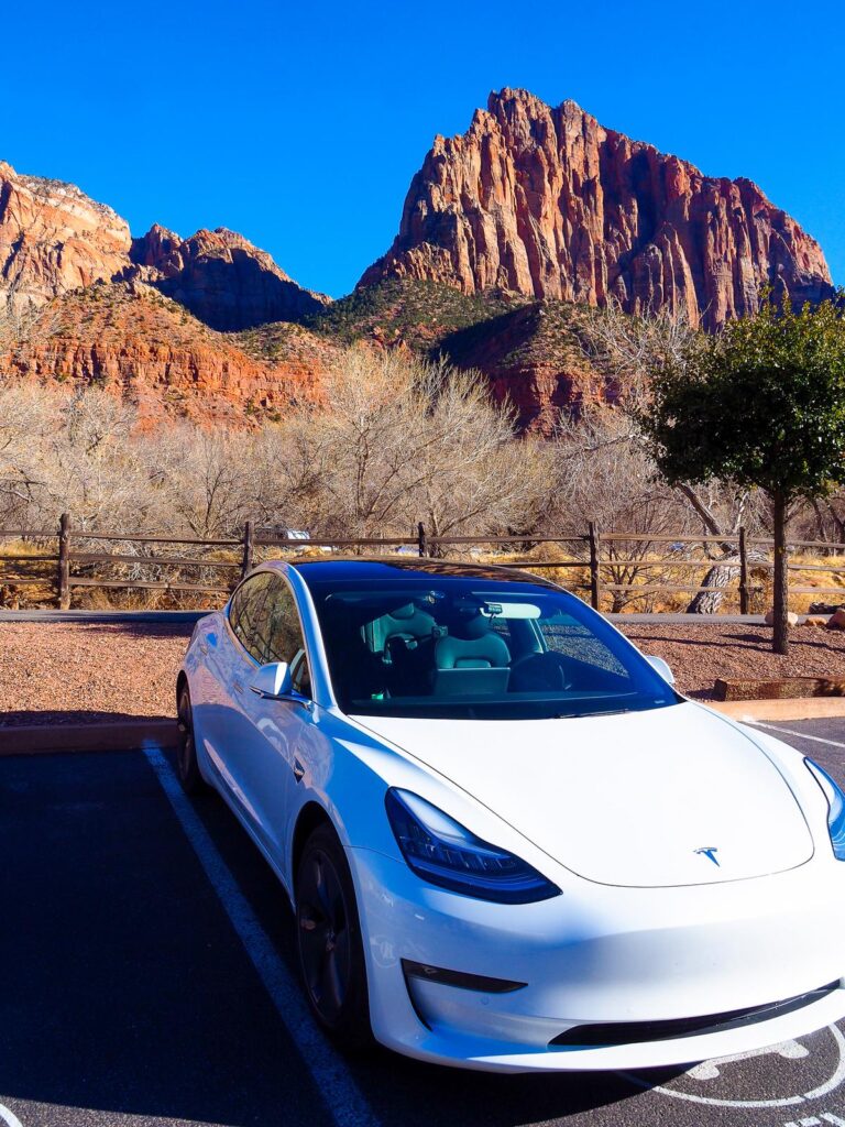 Alt-Text: White Tesla Model 3 charging against Zion National Park's red cliffs under blue skies