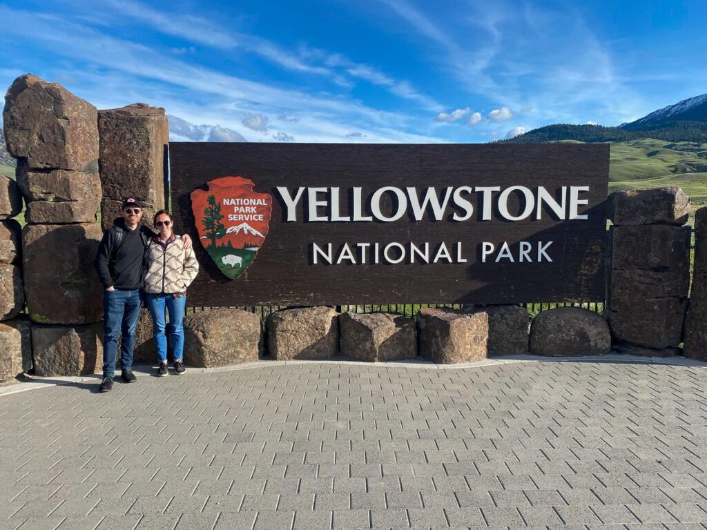 Alex and Kyle Kroeger of ViaTravelers at Yellowstone National Park entrance sign, with scenic mountain landscape.