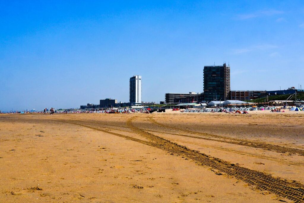 Lively Zandvoort beachfront with vacationers, coastal buildings, and tire tracks on sand in The Netherlands.